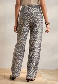 Mujer con vaqueros de tiro alto y pernera ancha con estampado de leopardo, sandalias negras de tacón bajo, camiseta blanca y pulseras en una muñeca, de pie sobre un suelo de baldosas.