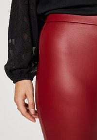 Høj talje, tætsiddende røde imitation læder leggings med en glat tekstur og minimal sømdetalje. Sort gennemsigtig bluseærme delvist synlig.