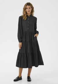 Woman with wavy hair wearing a long black buttoned dress with tiered skirt and black loafers, standing with one hand in pocket.