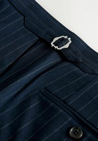 Navy pinstriped trousers made of textured fabric, featuring a silver metal clasp and a side pocket with a dark button.