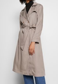 Trench coat beige con vita cinturata, chiusura a doppio petto con bottoni, ampie rever e maniche lunghe. Tessuto liscio, design fino al ginocchio.