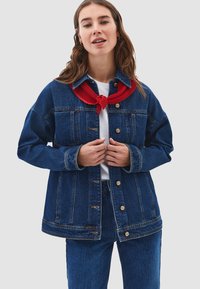 Kvinna med långt hår som bär en mörkblå denimjacka och byxor, vit skjorta och röd halsduk, står mot en ljus bakgrund.