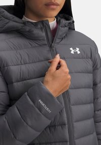 Šedá prešívaná bunda s kapucňou, ktorá má prešitý dizajn, zips a výšivku značky na rukáve a hrudi. Ľahký materiál.