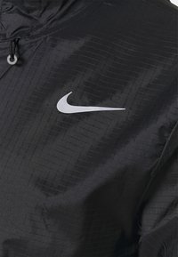 Veste légère noire en matériau brillant et texturé, avec un motif en grille et un logo Nike argenté réfléchissant sur la poitrine.