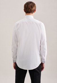 Camicia bianca a maniche lunghe con orlo curvo, polsini con bottoni e colletto classico. Tessuto dall'aspetto liscio; design minimalista.