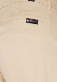 Beżowe bawełniane spodnie z tylną kieszenią z czarną metką z napisem „for all mankind”. Gładka tekstura i dopasowany krój.