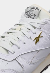 Baskets en cuir blanches Reebok avec lacets blancs, logo doré sur le côté et languette marquée affichant le nom Reebok et le symbole de la feuille d'érable.