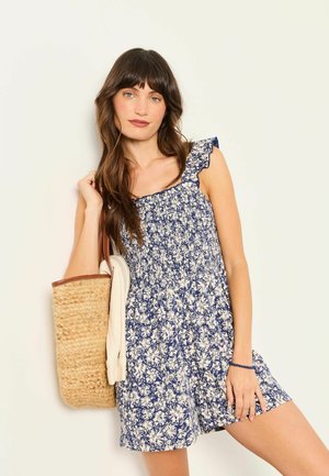 Woman with long brown hair wearing a blue floral sleeveless dress, carrying a woven straw tote bag with white fabric inside.