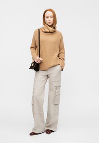 Pull lâche couleur chameau à col bénitier, associé à un pantalon cargo large beige avec poches latérales et détail zippé. Des chaussures marron complètent le look.