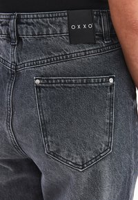 Jeans de mezclilla negra que presentan un bolsillo trasero con herrajes de metal y una etiqueta que muestra el nombre de la marca "OXXO". La tela tiene un acabado texturizado.