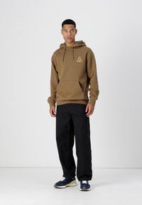 Brauner Kapuzenpullover mit einer frontalen Tasche und gelbem geometrischem Logo, kombiniert mit schwarzen lässigen Jeans und blauen Sneakers mit weißen Sohlen.