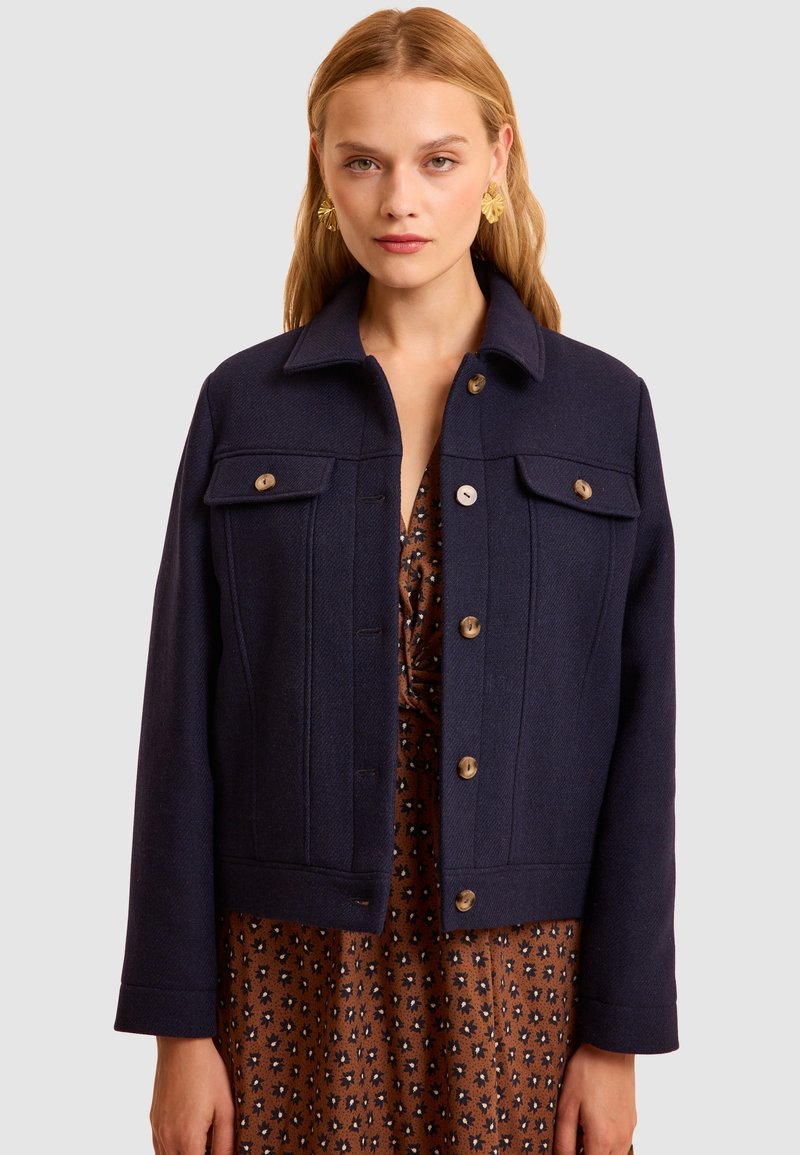 Giacca di lana blu navy con chiusura a bottone, due tasche sul petto e colletto. Presenta un tessuto texturizzato e un design corto.