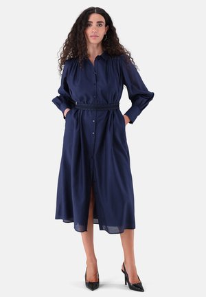 Frau mit langen lockigen Haaren, die ein marineblaues, langärmliges Midikleid mit Gürtel und schwarze High Heels trägt, steht mit den Händen in den Taschen.
