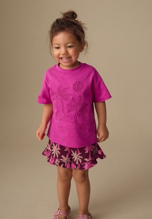 Ung pige smiler iført en pink skjorte med palme-tråd og matchende blomstrede flæse-short og sandaler på en beige baggrund.
