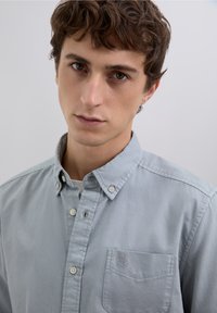 Lysegrå skjorte med knapper og syet brystlomme samt diskret logodetalje. Glat tekstur og klassisk kravedesign.