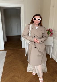 Femme portant un trench-coat beige, des lunettes de soleil blanches, une jupe en dentelle et des sandales blanches, debout à l'intérieur près d'une table blanche avec des fleurs roses.
