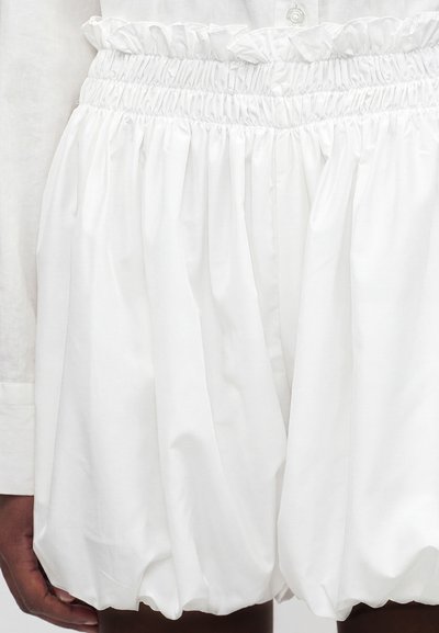 Short blanc taille haute en tissu froncé avec une ceinture élastique et un poignet de chemise blanc boutonné visible sur le côté gauche.