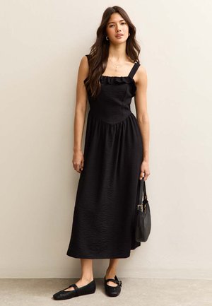 Femme en robe midi noire sans manches avec un col à volants tenant un sac à main noir, portant des chaussures plates noires, debout contre un mur beige.
