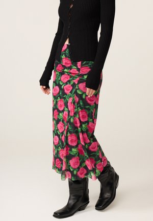 Vrouw draagt een zwarte gebuttonde cardigan, een lange bloemenrok met roze rozen en groene bladeren, en zwarte leren enkellaarzen.