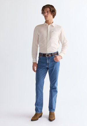 Homme portant une chemise crème à boutons, un jean bleu avec une ceinture marron, et des bottes en cuir marron, debout avec une main sur la hanche.