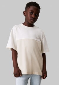 Camiseta de manga corta en algodón beige y blanco roto, con un diseño de bloques de color y un pequeño logo en el pecho. Textura suave.