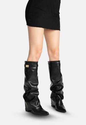 Ben iført en sort tekstureret mininederdel og knæhøje, løstsiddende læderstøvler med spidse tæer og små blokhæle.