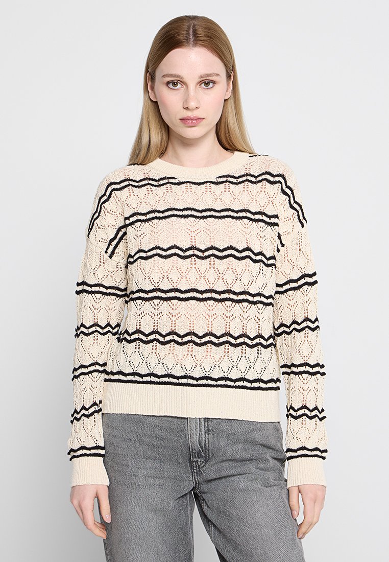 Jonge vrouw met steil blond haar, gekleed in een crèmekleurige gebreide trui met zwarte zigzagstrepen en grijze spijkerbroek, staande tegen een witte achtergrond.