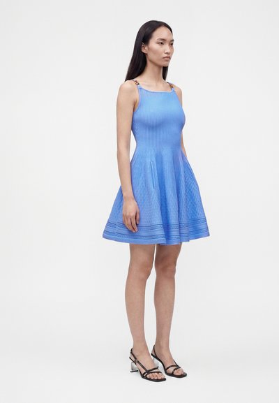 Femme aux longs cheveux foncés portant une robe tricotée bleue sans manches et des talons noirs à brides, debout devant un fond blanc uni.