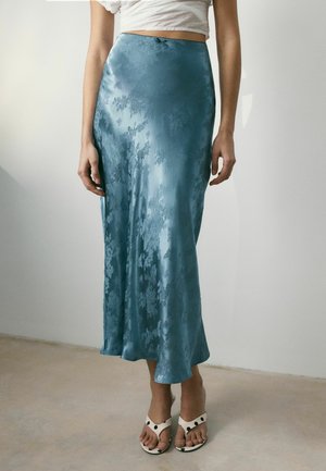 Woman wearing a blue floral satin midi skirt, white cropped top, and white sandals with black polka dots standing on a beige floor.