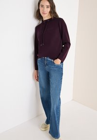 Burgunderfarbener Kapuzenpulli mit Kordelzug und geprägtem Logo, kombiniert mit hellblauen weiten Jeans und beigen Sneakers vor einem neutralen Hintergrund.