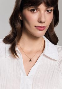 Złoty naszyjnik z łańcuchem z wisiorkiem w ciemnym okrągłym kształcie oraz dekoracyjnymi elementami. Nosi się go na teksturowanej białej bluzce.