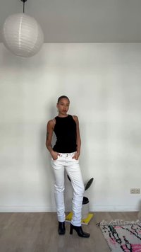 Egy személy beltérben áll, fekete ujjatlan felsőt, fehér nadrágot és fekete, sarkú csizmát visel, mellette papírlámpás és növény lóg.