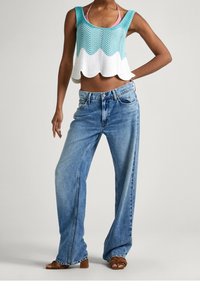 Kvinna iklädd lösa blå jeans, öppna bruna sandaler och en ärmlös turkos och vit virkade crop top.