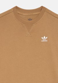 Bež pulover s krožnim izrezom, ki vsebuje šivano v-detail na vratu in belo Adidasovo logo na levem prsnem delu. Mehka, gladka tekstura.