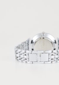 Silberne Metalluhr mit einem Gliedband, runder Gehäuseform, polierter Oberfläche und zentralem Verschluss. Minimalistisches Design ohne sichtbare Ziffernblattdetails.