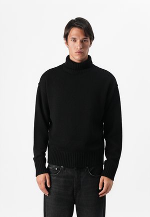 Črna turtleneck pulover z ohlapen fit, rebrastimi manšetami in robom, narejen iz teksturirane pletene tkanine. V kombinaciji z temnimi kavbojkami.