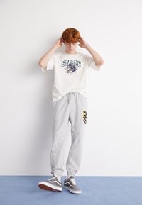 Hvid kortærmet t-shirt med "STAPLE" grafik og tigermotiv. Grå sweatpants med gul og sort print. Grå sneakers med hvide accenter.
