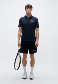 Mornarsko modra polo majica z logotipom, združena s črnimi kratkimi hlačami. Model drži teniški lopar, nosi bele nogavice in superge. Minimalističen dizajn.