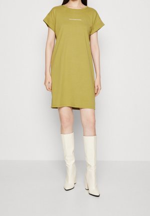 Olivgrünes Baumwoll-T-Shirt-Kleid mit kurzen Ärmeln und rundem Ausschnitt, mit Text auf der Brust. Kombiniert mit kniehohen weißen Stiefeln.