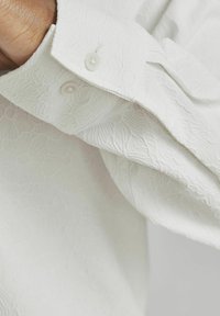 Witte langekmouwen shirt met een textuur van een bloemenpatroon, voorzien van twee knopen op de manchet, gemaakt van een lichtgewicht stof.