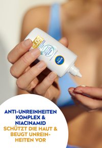 Sonnenschutzmittel-Flasche mit weißem Kunststoffkörper und spitzem Ende. Blaue und goldene Etikettierung, die SPF 50+ anzeigt. Person, die Creme auf ihren Finger gibt.