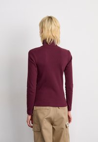 Persona con capelli biondi corti che indossa un dolcevita bordeaux e pantaloni cargo beige, in piedi di fronte a uno sfondo bianco semplice.