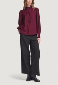 Blusa bordeaux con fronte arricciato, chiusura a bottoni e maniche lunghe abbinata a pantaloni neri a gamba larga e stivaletti.