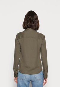 Camisa verde oliva de manga larga y cuello. Presenta una textura suave y puños con botones. La parte trasera tiene un diseño de yugo y un dobladillo curvado.