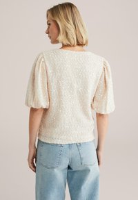 Žena s krátkými blond vlasy, oblečená v bílém topu se třpytkami a nabíranými rukávy, a světle modrých džínách s vysokým pasem, stojící zády k béžovému pozadí.