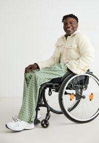 Witte fleecjas, groene geblokte broek en witte sneakers; zittend in een zwarte rolstoel met oranje accenten en zilveren wielen op een lichte achtergrond.