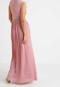 Lange, geplooide jurk van zachtroze stof met een aansluitende lijfje en hoge halslijn. Heeft een ritssluiting aan de achterkant en een uitlopende rok.