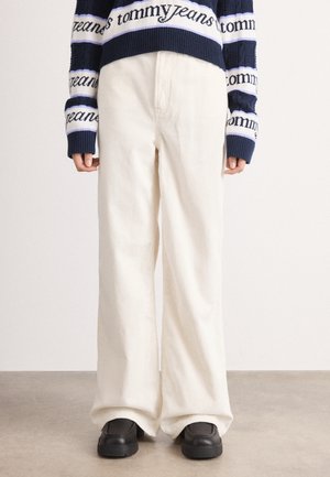 Person trägt cremefarbene weite Cordhosen, schwarze klobige Loafer und einen dunkelblauen Pullover mit der Aufschrift "tommy jeans" auf den Ärmeln und am Saum.