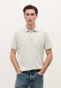Helles graues Poloshirt mit strukturierter Oberfläche, kurzen Ärmeln und einer Zwei-Knopf-Leiste, kombiniert mit blauen Jeans und einem schwarzen Gürtel.