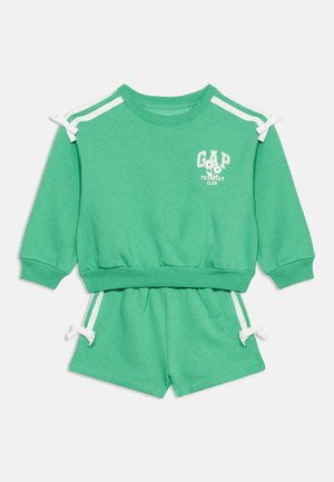 Completo verde da bambini composto da top a maniche lunghe e pantaloncini, con strisce bianche sulle spalle, fiocchi e scritta "GAP Friendship Club" con fiori.